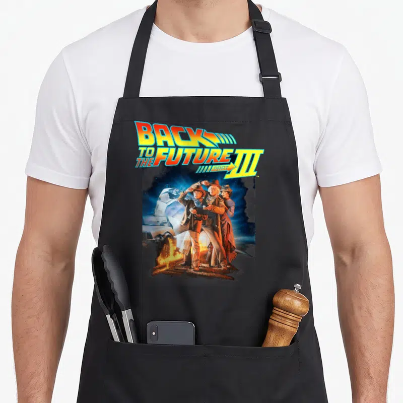 Three movie poster apron - Stacked Back to the Future Part III text above photo of three characters standing on train — shown on a black cotton canvas apron against a white background