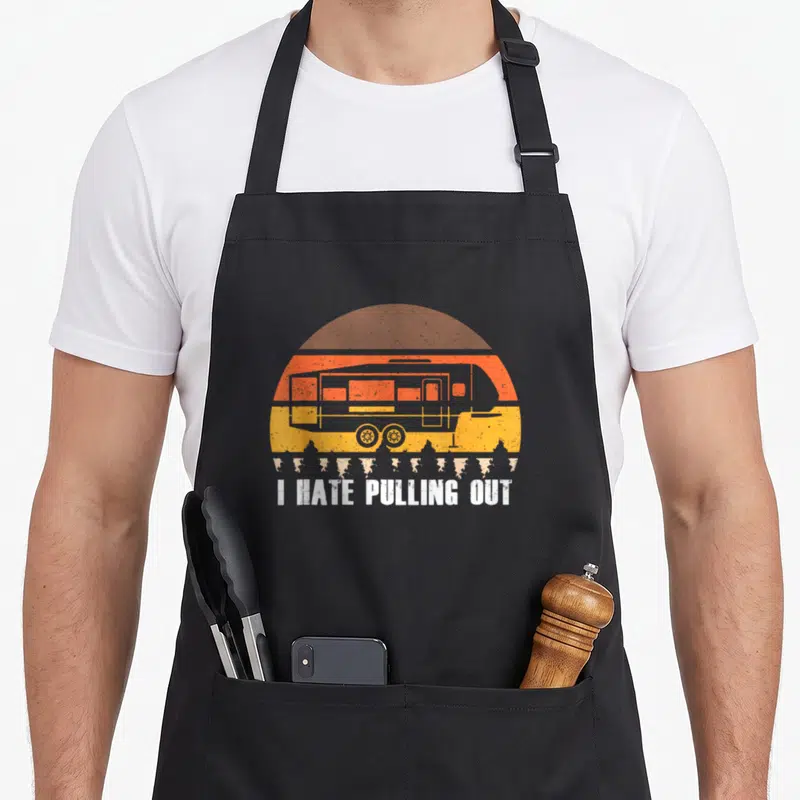 Hate pulling camping apron - Retro camper trailer in orange and yellow sun with white text I HATE PULLING OUT — shown on a black cotton canvas apron against a white background