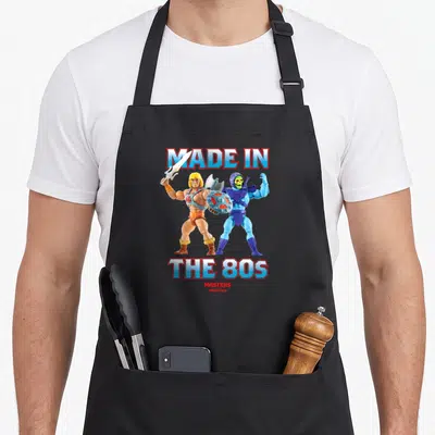 Funny 80s apron - He-Man and Skeletor standing back-to-back under Made in The 80s text — shown on a black cotton canvas apron against a white background
