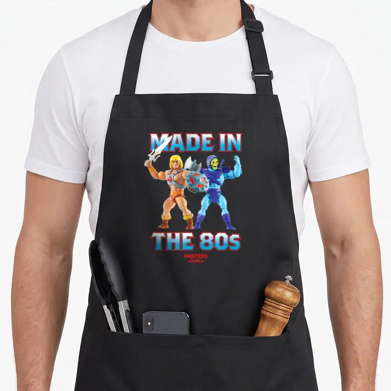 Funny 80s apron - He-Man and Skeletor standing back-to-back under Made in The 80s text — shown on a black cotton canvas apron against a white background
