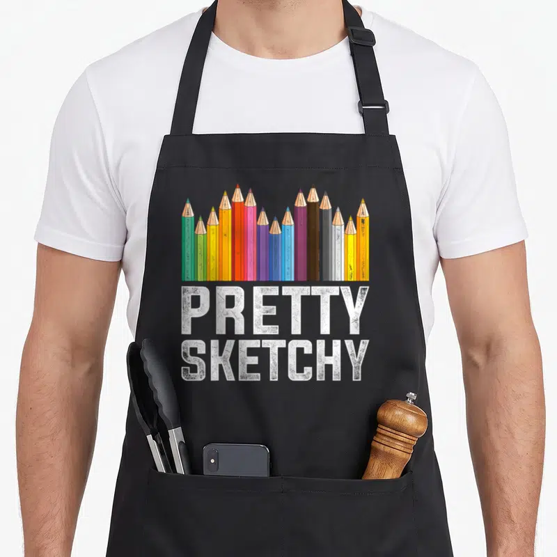 Artist pretty apron - Row of colored pencils above stacked white text reading pretty sketchy — shown on a black cotton canvas apron against a white background