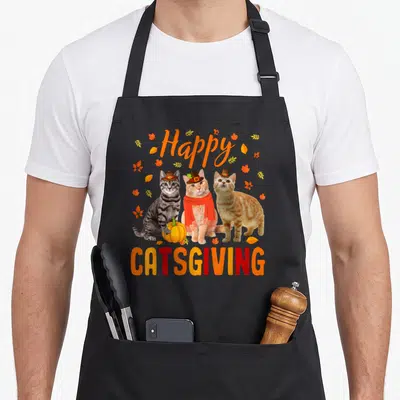 Catsgiving pilgrim apron - Three cats in pilgrim hats with a pumpkin and autumn leaves under Happy Catsgiving text — shown on a black cotton canvas apron against a white background