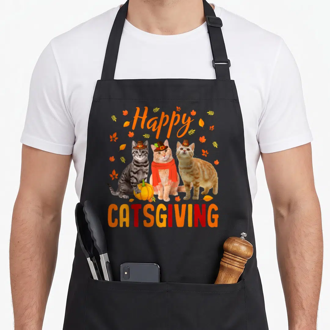 Catsgiving pilgrim apron - Three cats in pilgrim hats with a pumpkin and autumn leaves under Happy Catsgiving text — shown on a black cotton canvas apron against a white background