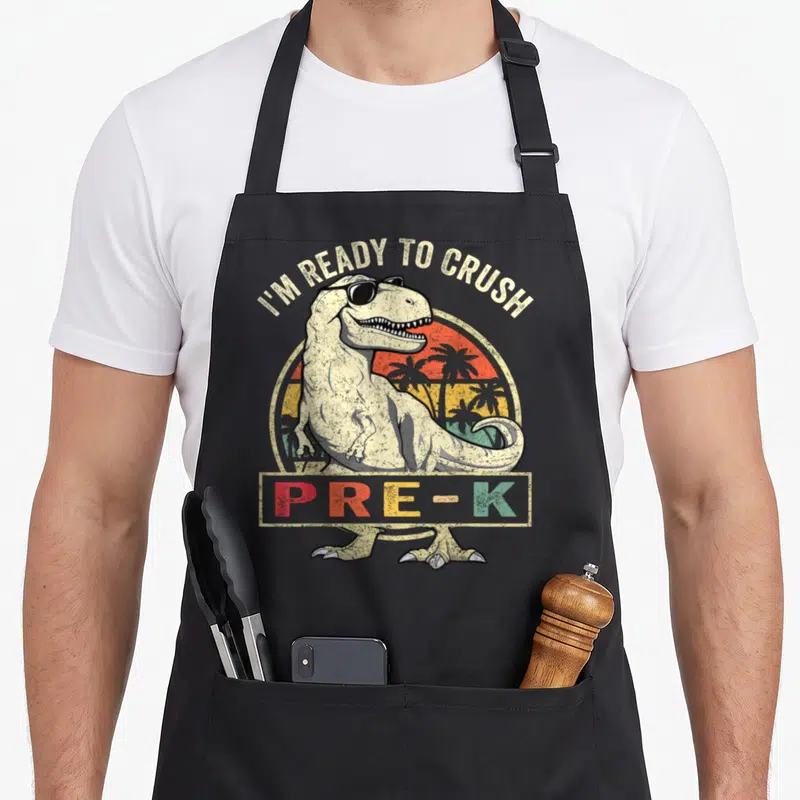 Crush pre dinosaur apron - Retro T-Rex with sunglasses and I'M READY TO CRUSH PRE-K text on black apron — shown on a black cotton canvas apron against a white background