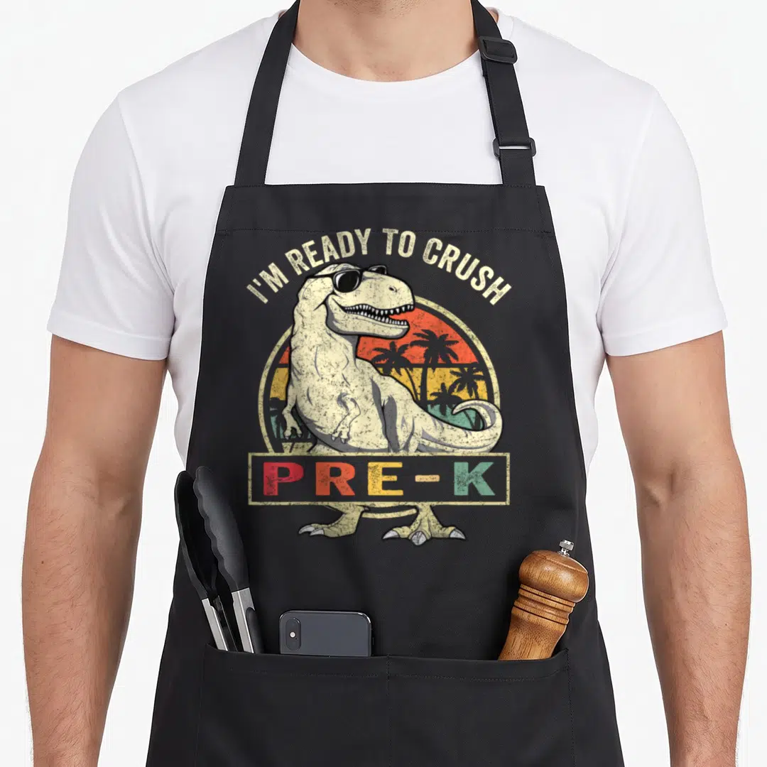Crush pre dinosaur apron - Retro T-Rex with sunglasses and I'M READY TO CRUSH PRE-K text on black apron — shown on a black cotton canvas apron against a white background