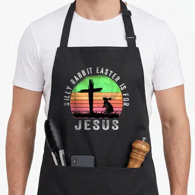 Christian rabbit apron - Silly Rabbit Easter is for Jesus text over distressed sunset circle with black cross — shown on a black cotton canvas apron against a white background