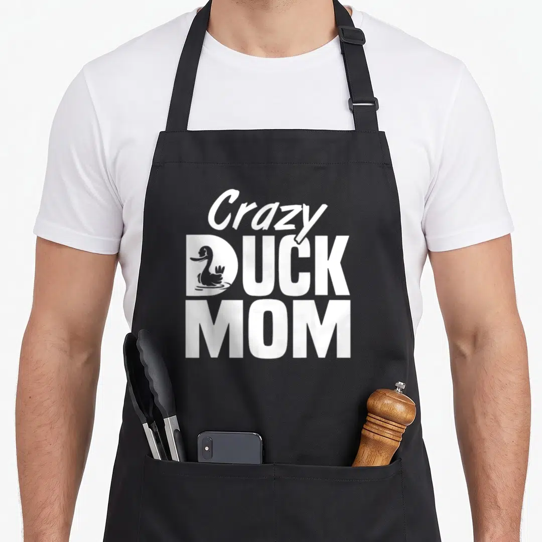 Duck mother's apron - Stacked white text Crazy DUCK MOM with duck inside D, no color fill — shown on a black cotton canvas apron against a white background
