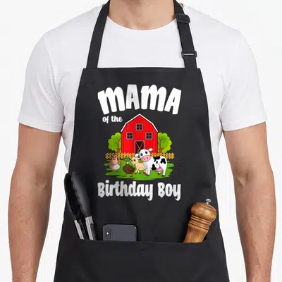 Mama farm aprons - Cartoon red barn with cow duck goat chicken and MAMA of the Birthday Boy text — shown on a black cotton canvas apron against a white background