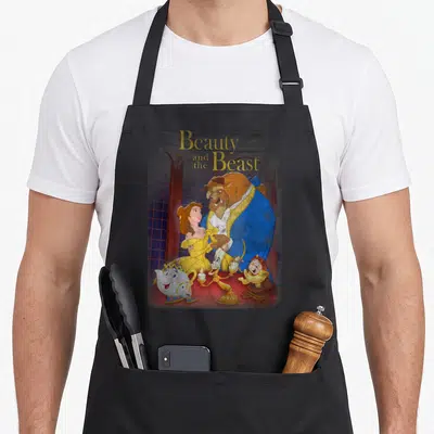 Shot distressed apron - Beauty and the Beast title above Beast Belle and talking objects on red table with arched — shown on a black cotton canvas apron against a white background