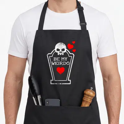 Gothic valentine's day apron - White coffin outline with skull, red hearts, and distressed text reading BE MY WEIRDO — shown on a black cotton canvas apron against a white background