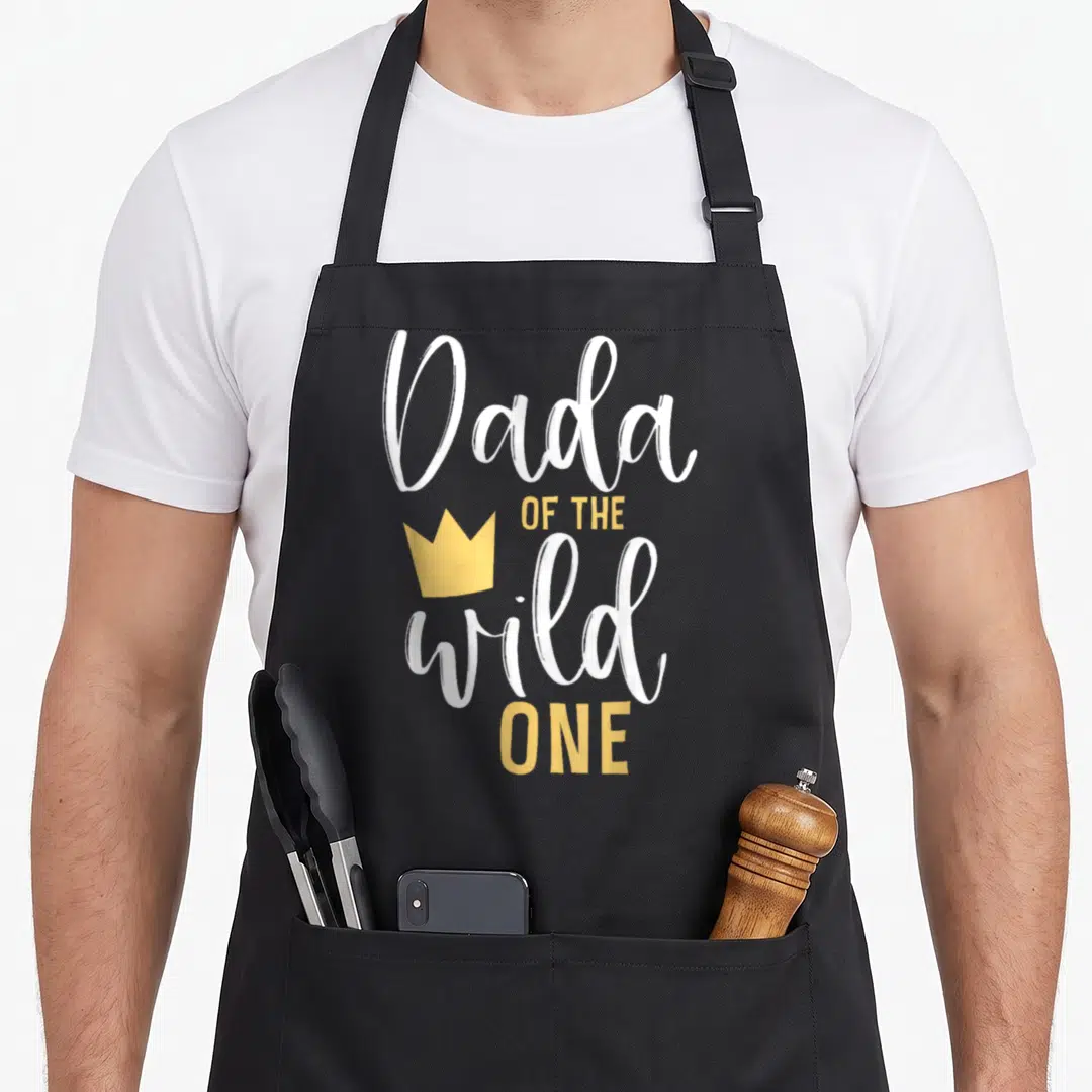 Dada birthday apron - White and yellow text reading Dada OF THE wild ONE with a crown icon — shown on a black cotton canvas apron against a white background