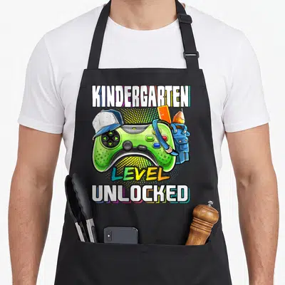 Kindergarten level video game apron - Green game controller with hat, orange ruler, pencil, blue overalls, and stacked — shown on a black cotton canvas apron against a white background