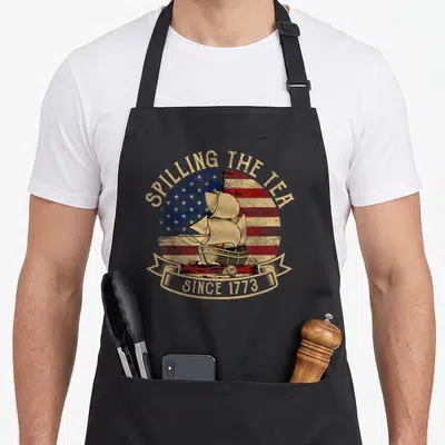 Spilling tea apron - Retro sailing ship on circular flag with text SPILLING THE TEA SINCE 1773 — shown on a black cotton canvas apron against a white background