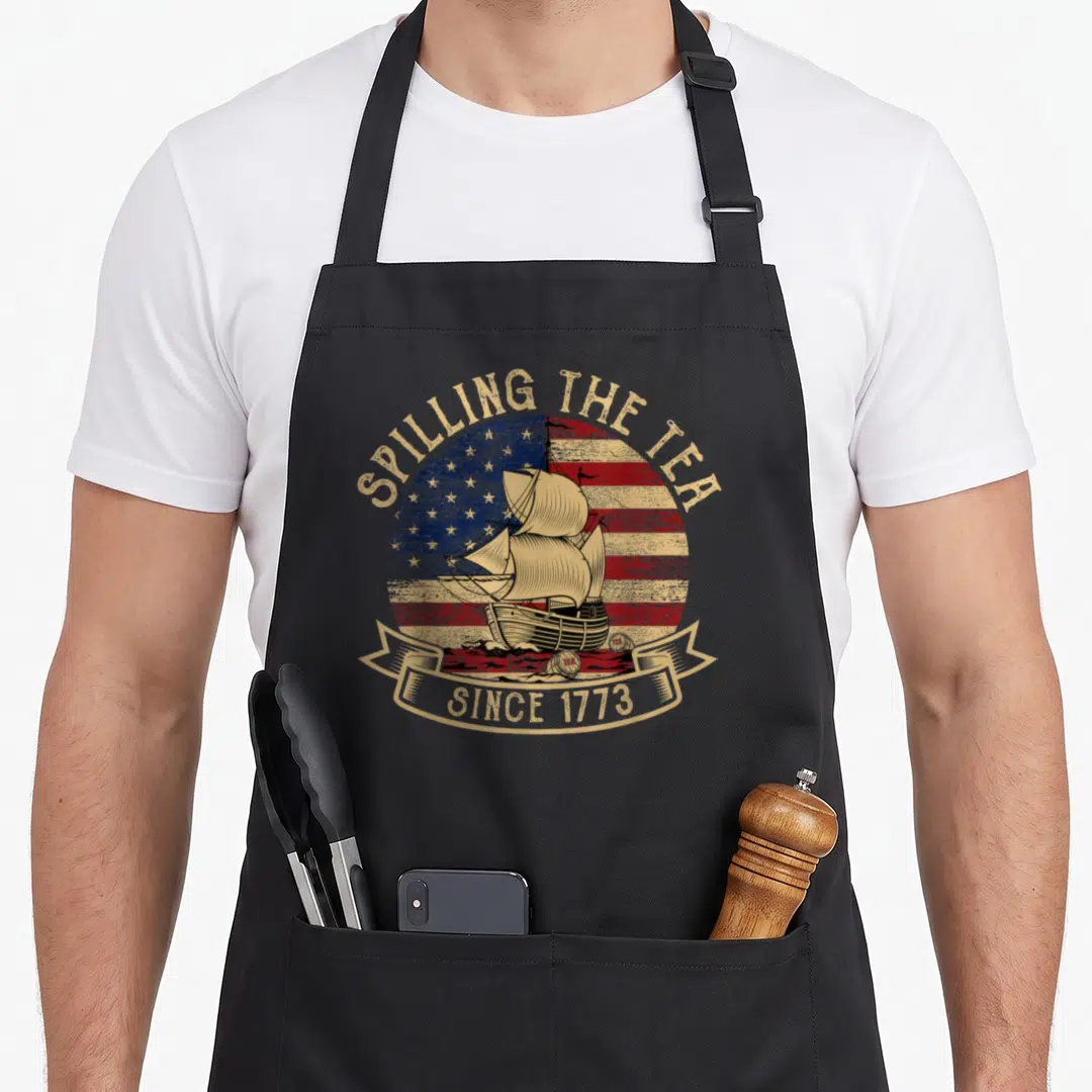 Spilling tea apron - Retro sailing ship on circular flag with text SPILLING THE TEA SINCE 1773 — shown on a black cotton canvas apron against a white background