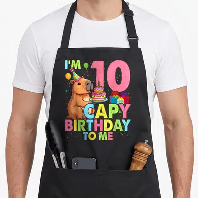 Bday capybara birthday apron - Cartoon capybara holding cake next to pink 10 and colorful birthday text — shown on a black cotton canvas apron against a white background