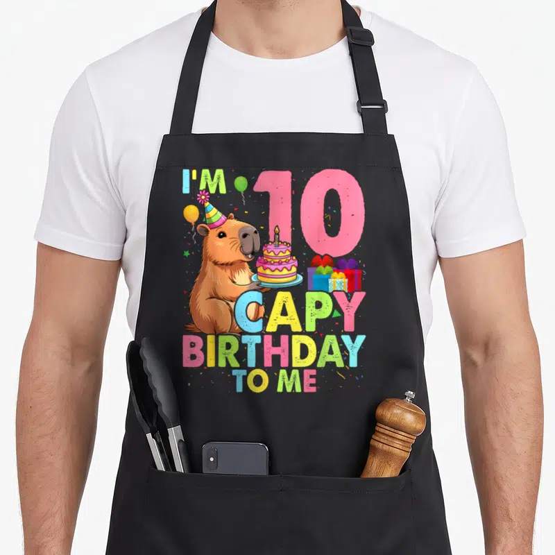 Bday capybara birthday apron - Cartoon capybara holding cake next to pink 10 and colorful birthday text — shown on a black cotton canvas apron against a white background