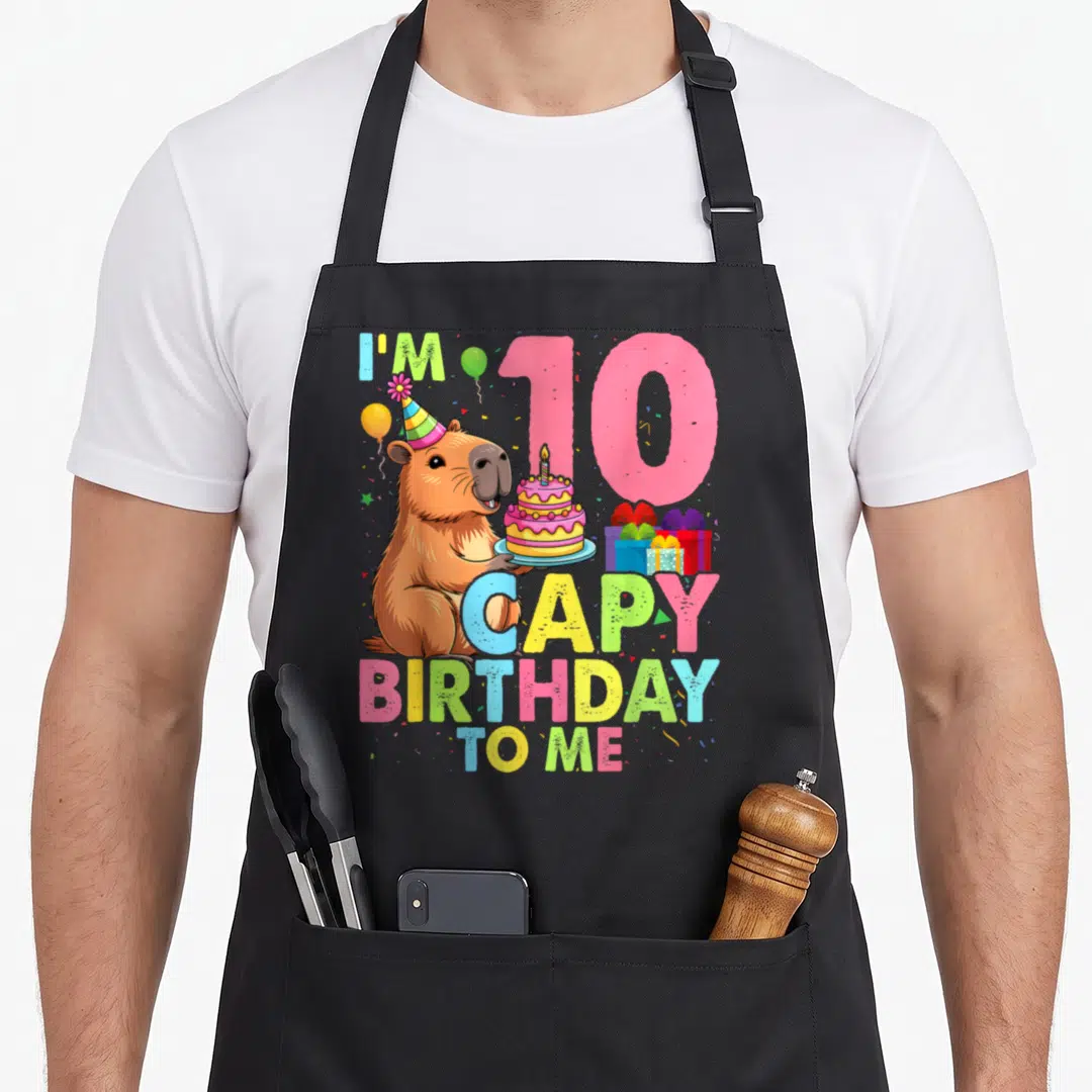 Bday capybara birthday apron - Cartoon capybara holding cake next to pink 10 and colorful birthday text — shown on a black cotton canvas apron against a white background