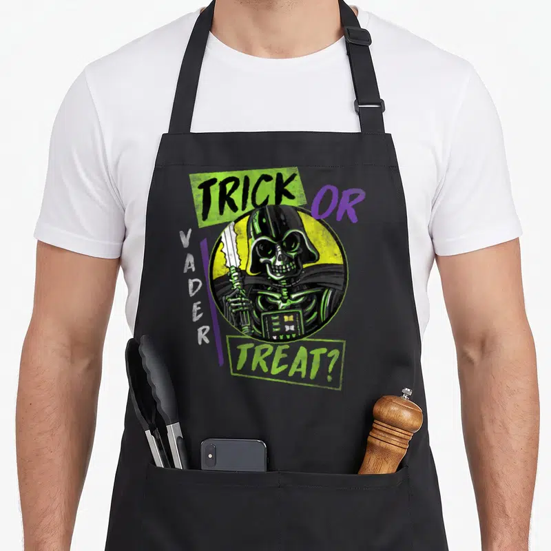 Vintage trick halloween apron - Skeletal Darth Vader in yellow circle with TRICK OR VADER TREAT? text in lime green — shown on a black cotton canvas apron against a white background