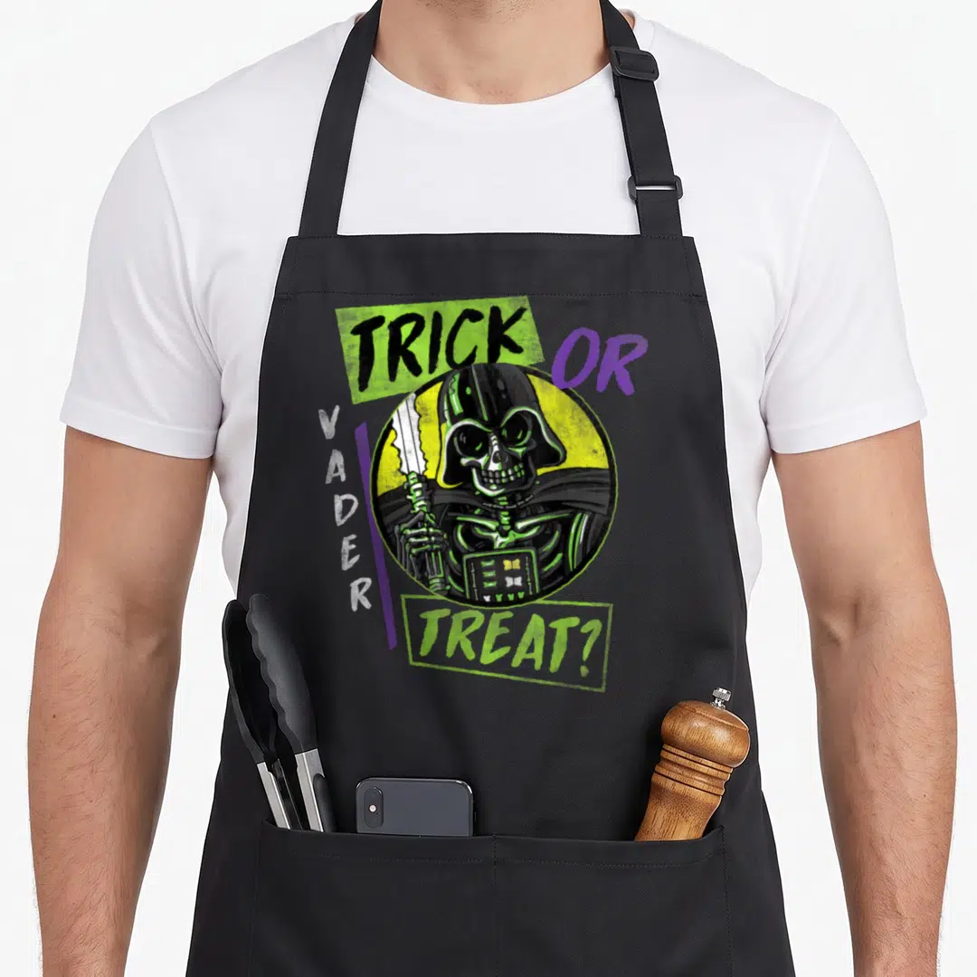 Vintage trick halloween apron - Skeletal Darth Vader in yellow circle with TRICK OR VADER TREAT? text in lime green — shown on a black cotton canvas apron against a white background