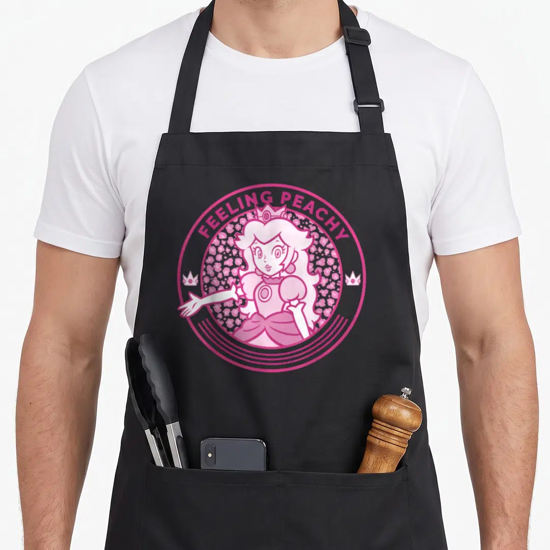 Princess cheetah apron - Cartoon woman inside pink circle with hearts and two crowns, text reading Feeling Peachy — shown on a black cotton canvas apron against a white background