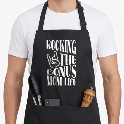 Rocking bonus mother's day aprons - White stacked text ROCKING THE BONUS MOM LIFE with rock-on hand symbol next — shown on a black cotton canvas apron against a white background