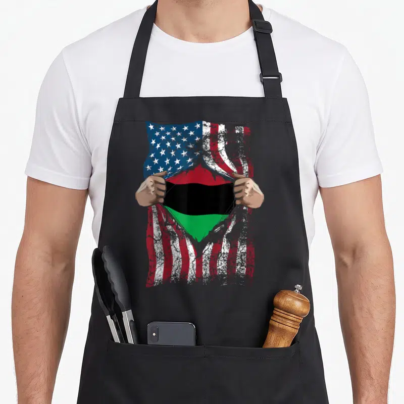 Pan american flag apron - Distressed USA flag torn open by two hands revealing red, black, and green striped flag — shown on a black cotton canvas apron against a white background