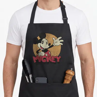 Distressed best friend aprons - Retro Mickey Mouse waving in front of tan circle with red MICKEY text overlay — shown on a black cotton canvas apron against a white background