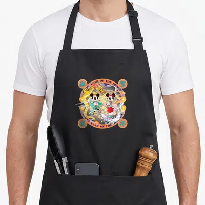 Funny lunar dragon apron - Mickey and Minnie Mouse surrounded by a dragon with HAPPY YEAR OF THE DRAGON text arcs — shown on a black cotton canvas apron against a white background