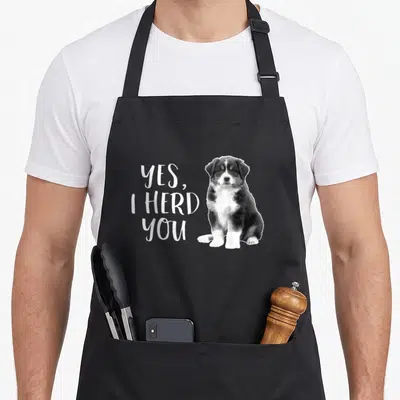 Shepherd australian apron - Yes I Herd You text next to a sitting black white and brown puppy photo — shown on a black cotton canvas apron against a white background