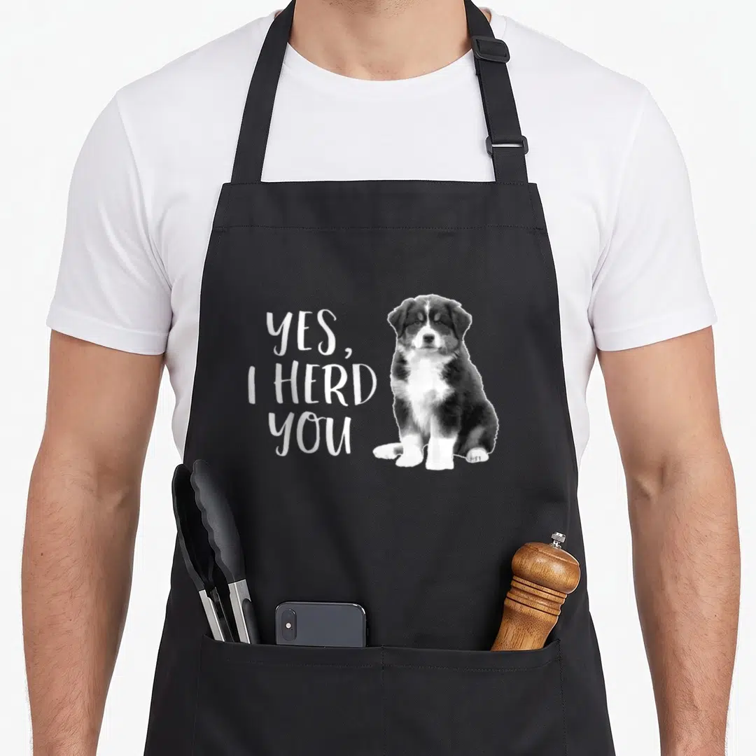 Shepherd australian apron - Yes I Herd You text next to a sitting black white and brown puppy photo — shown on a black cotton canvas apron against a white background