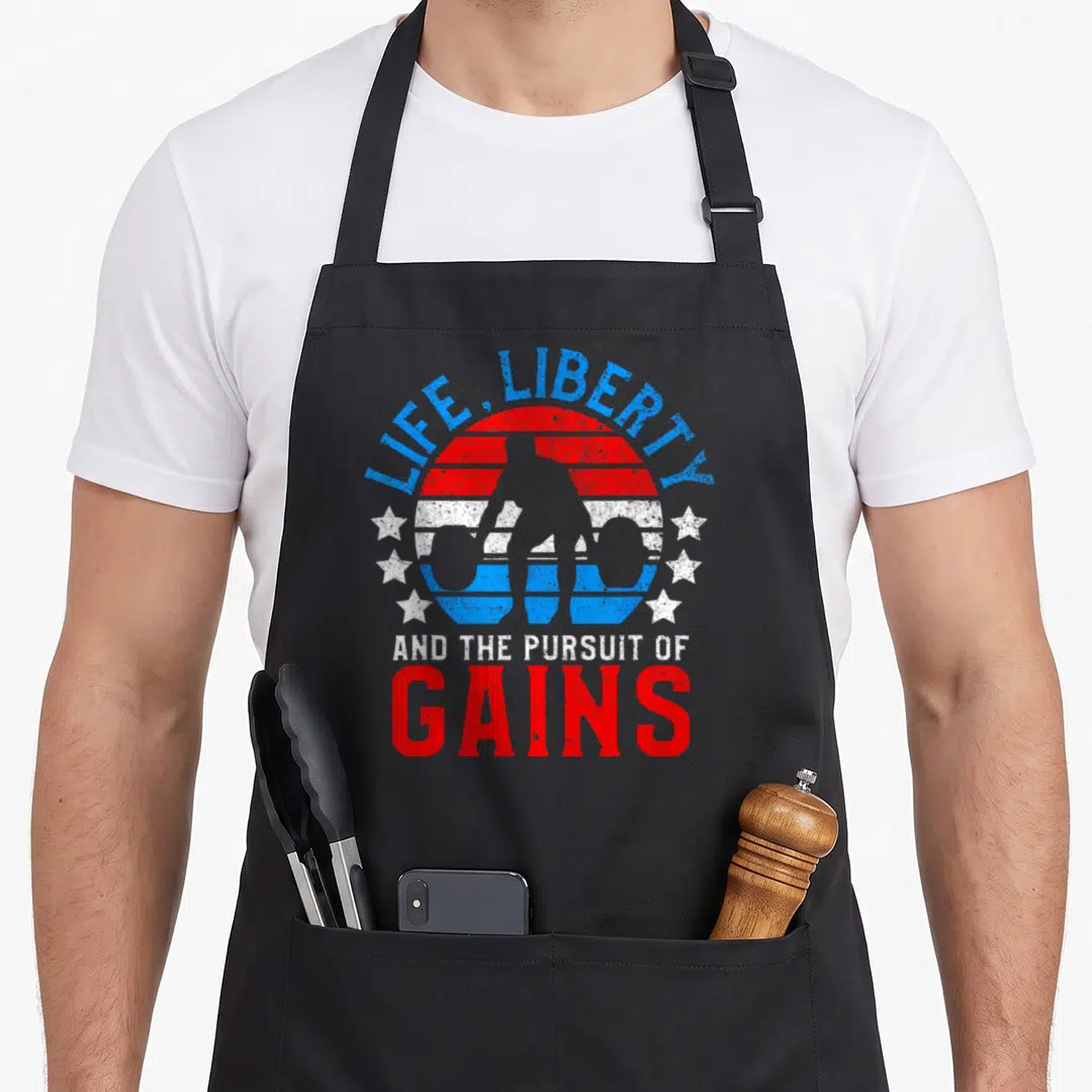 Gain liberty apron - Retro weightlifter silhouette in red white and blue striped circle with stars and pursuit of gains — shown on a black cotton canvas apron against a white background