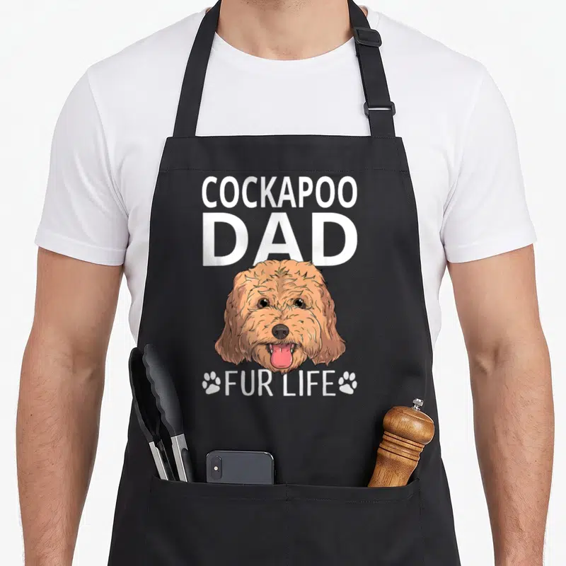 Father cockapoo apron - Cartoon white and tan dog head with COCKAPOO DAD text and FUR LIFE flanked by paw prints — shown on a black cotton canvas apron against a white background