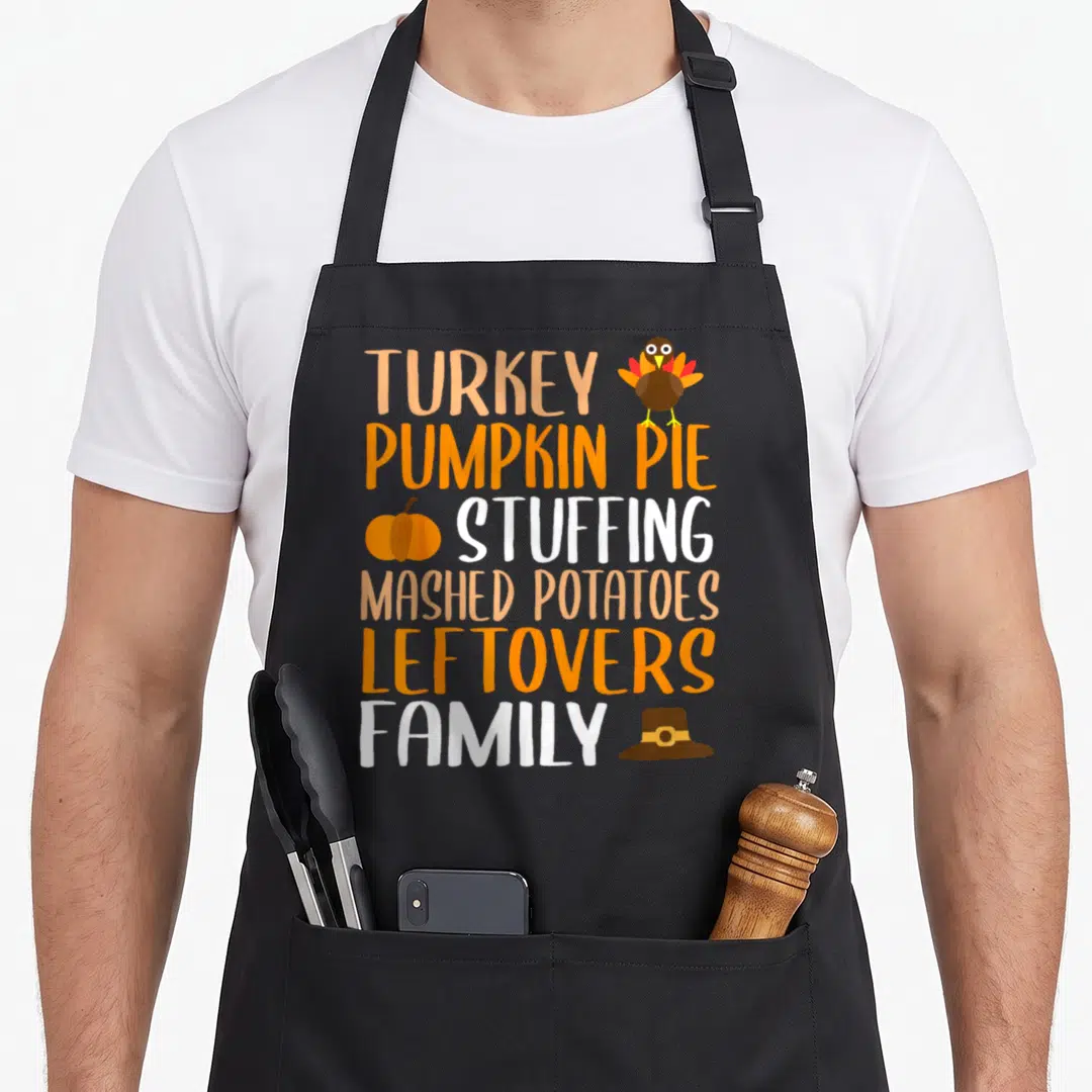 Pie thanksgiving aprons - Stacked text listing turkey, pie, stuffing, potatoes, leftovers, family with cartoon turkey — shown on a black cotton canvas apron against a white background