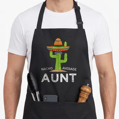 Nacho aunt apron - Cactus in sombrero with NACHO AVERAGE AUNT text in green orange white — shown on a black cotton canvas apron against a white background