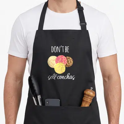 Self spanish aprons - Four cartoon conchas in pink, yellow, brown, and white with text Don't Be self conchas — shown on a black cotton canvas apron against a white background