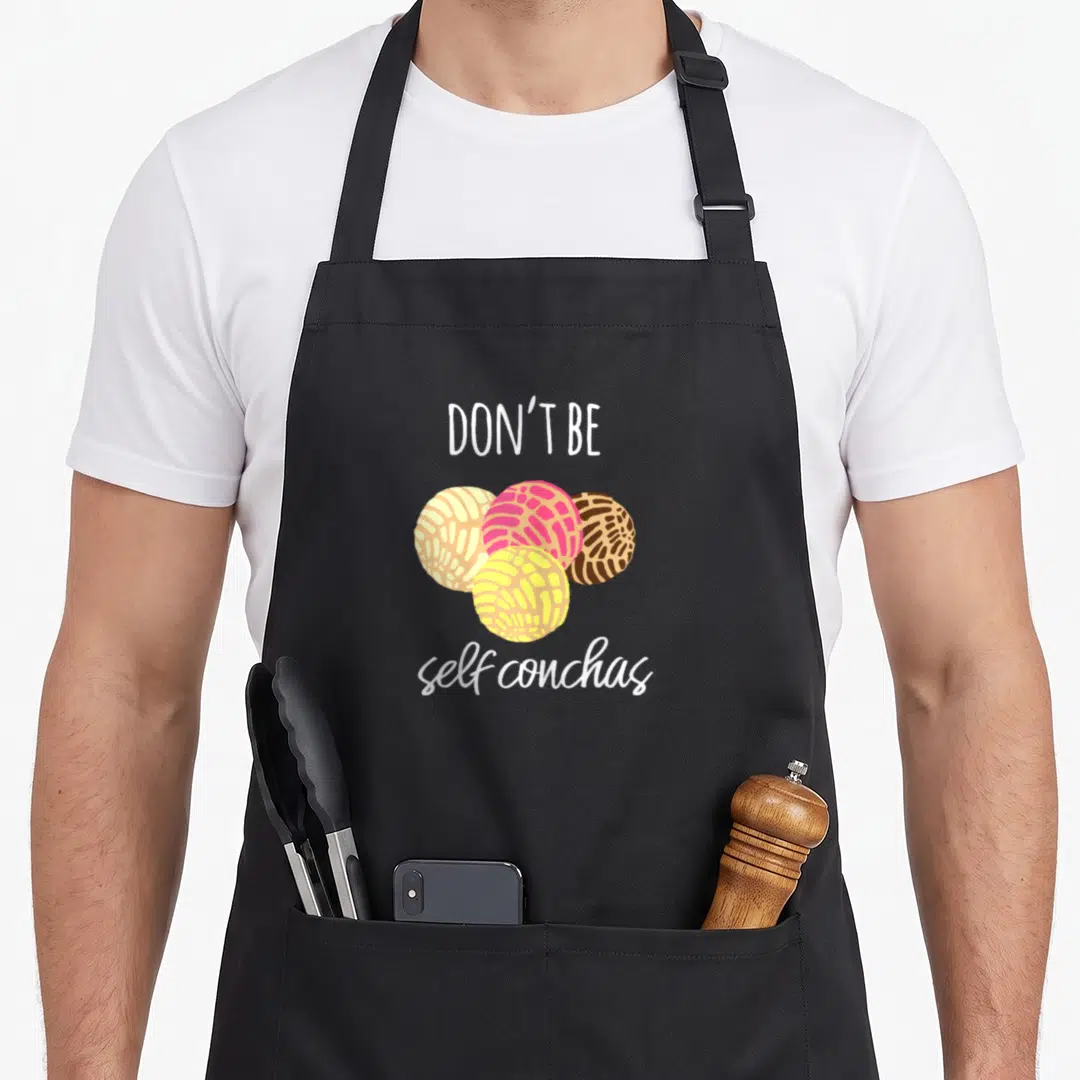 Self spanish aprons - Four cartoon conchas in pink, yellow, brown, and white with text Don't Be self conchas — shown on a black cotton canvas apron against a white background