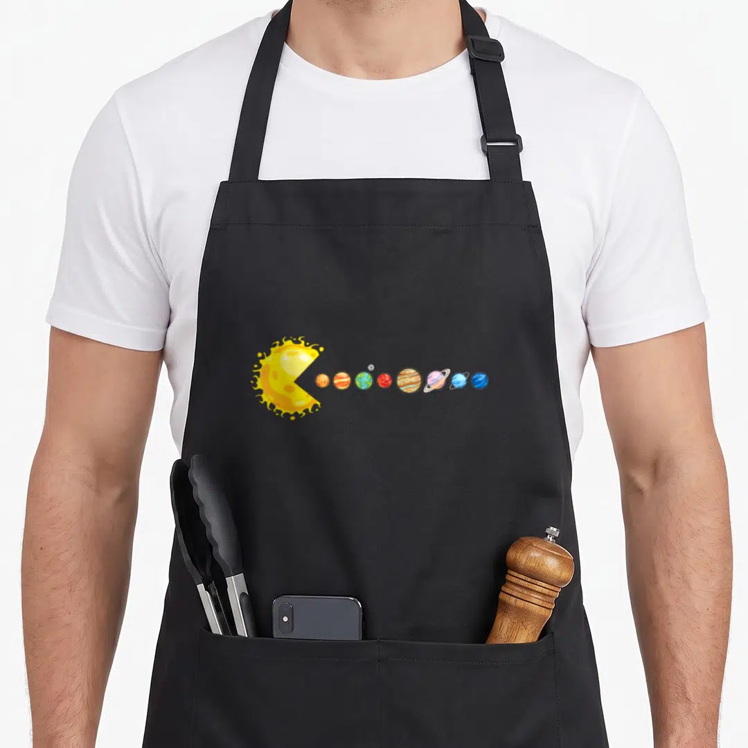 Planetary space planets apron - Cartoon yellow sun with open mouth facing a row of multi-colored planets — shown on a black cotton canvas apron against a white background