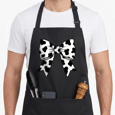 Cowprint cow apron - Black and white cow print cartoon bow graphic on a black bib apron — shown on a black cotton canvas apron against a white background