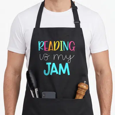 Read teacher apron - Stacked colorful text reading Reading is my Jam in blue pink yellow green and white fonts — shown on a black cotton canvas apron against a white background