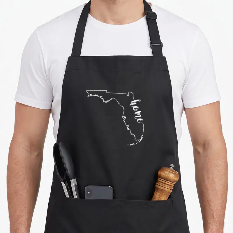 Floridian florida apron - White Florida state outline with home text running vertically along the eastern border — shown on a black cotton canvas apron against a white background