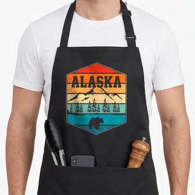 Landscape bear apron - Retro hexagon with ALASKA text, layered mountains, trees, and bear in red orange yellow teal — shown on a black cotton canvas apron against a white background