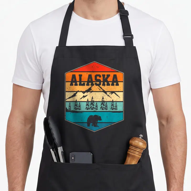 Landscape bear apron - Retro hexagon with ALASKA text, layered mountains, trees, and bear in red orange yellow teal — shown on a black cotton canvas apron against a white background