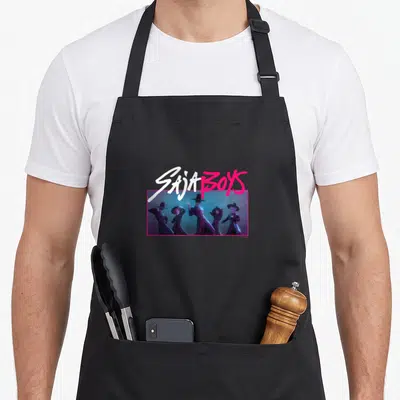 Saja novelty apron - SAJA BOYS text over pink frame with five men standing in row, photo style — shown on a black cotton canvas apron against a white background