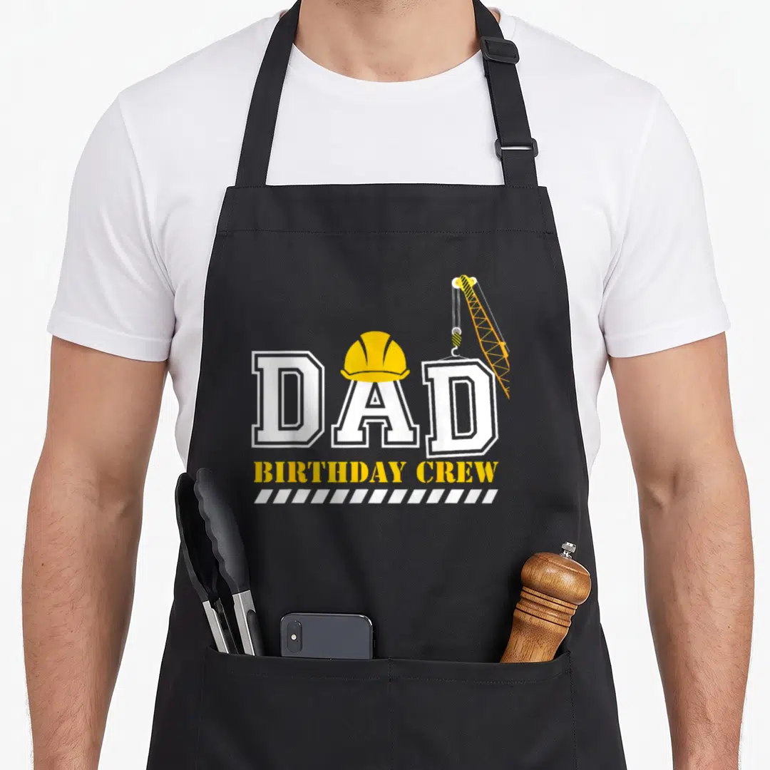 Birthday construction apron - DAD BIRTHDAY CREW text with yellow hard hat, crane hook, and hazard stripes — shown on a black cotton canvas apron against a white background