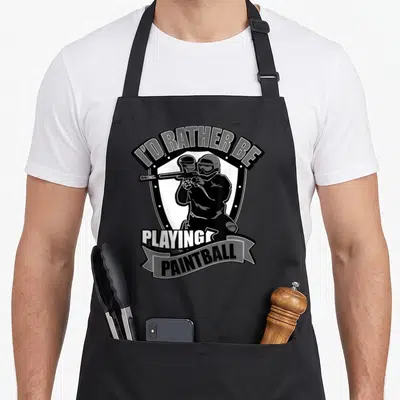 Balling paint apron - Shield graphic of paintball player holding marker with arching text I'D RATHER BE PLAYING — shown on a black cotton canvas apron against a white background
