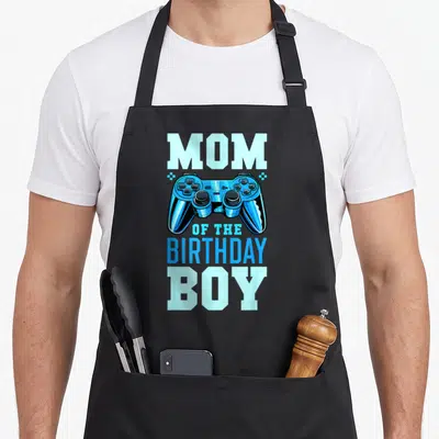 Retro video birthday apron - Blue game controller between MOM and OF THE BIRTHDAY BOY text flanked by two stars — shown on a black cotton canvas apron against a white background