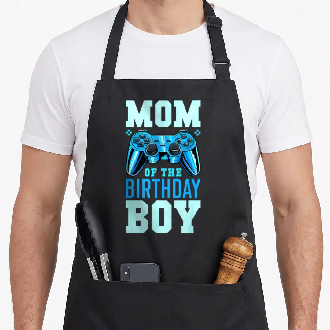 Retro video birthday apron - Blue game controller between MOM and OF THE BIRTHDAY BOY text flanked by two stars — shown on a black cotton canvas apron against a white background