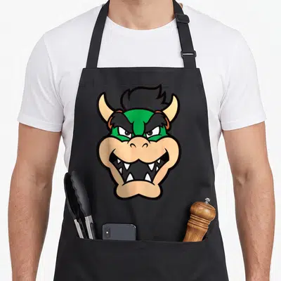 Chest holiday apron - Cartoon monster head with green skin and tan horns centered on apron — shown on a black cotton canvas apron against a white background