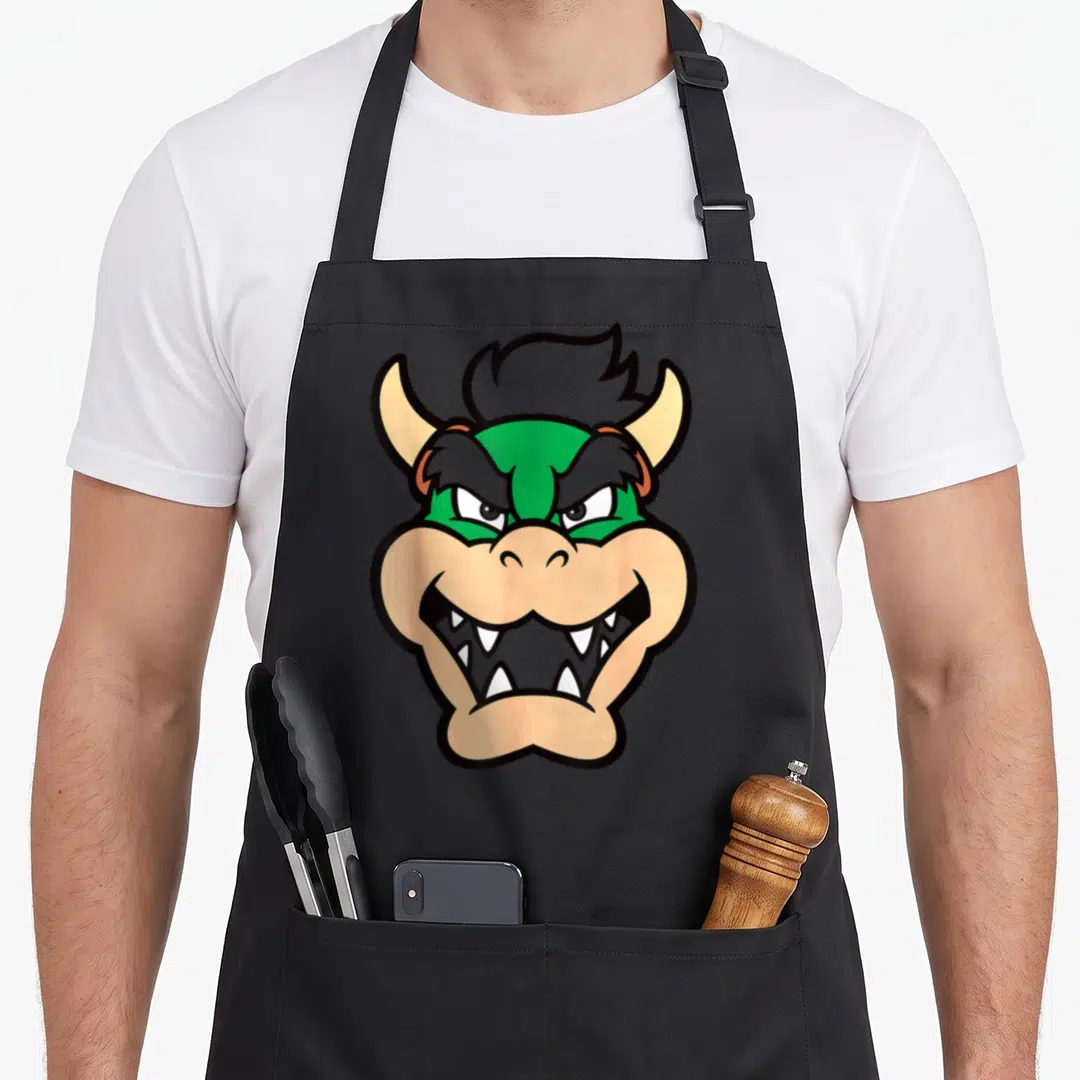 Chest holiday apron - Cartoon monster head with green skin and tan horns centered on apron — shown on a black cotton canvas apron against a white background