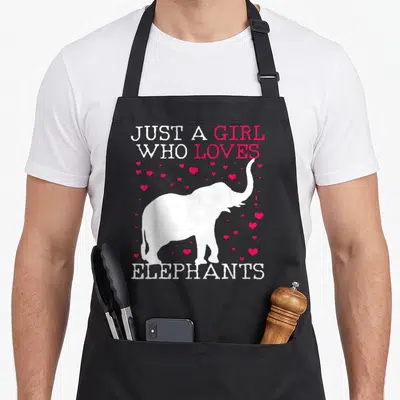 Cute elephant print apron - White elephant silhouette surrounded by pink hearts and text reading JUST A GIRL WHO LOVES — shown on a black cotton canvas apron against a white background
